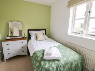 A bedroom with a single bed with white sheets and green blanket next to a window and a white chest of drawers with a mirror and clock at Rosemead in Moreton-In-Marsh