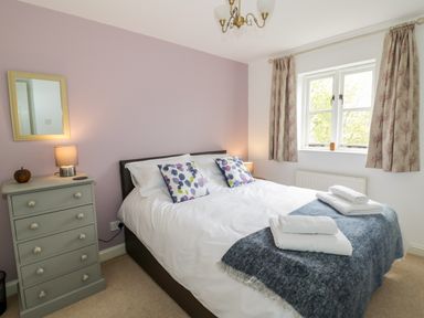 A bedroom with a double bed with white bedding and patterned pillows next to a window with curtains and a chest of drawers with a lamp and mirror at Rosemead in Moreton-In-Marsh