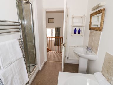 A bathroom with a shower, towel rack, pedestal sink, mirror, wall shelf with towels and bottles at Rosemead in Moreton-In-Marsh