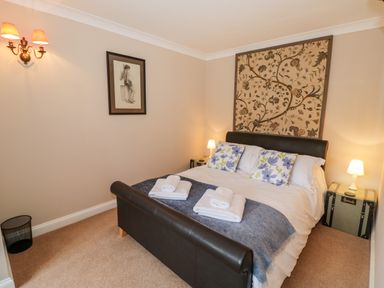 A bedroom with a double bed with floral pillows two folded towels floral wall hanging and bedside lamps at Rosemead in Moreton-In-Marsh