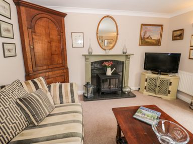 A living room with a striped sofa a wooden cabinet a fireplace with an oval mirror a TV on a corner stand and a wooden coffee table with a book at Rosemead in Moreton-In-Marsh