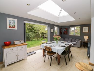 A dining area with a table and chairs next to a gray sofa and an open door to a garden at Rosemead in Moreton-In-Marsh