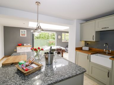 A kitchen with a granite island countertop holding a tray with food items and a wine bottle in a cooler next to a wooden cutting board at Rosemead in Moreton-In-Marsh