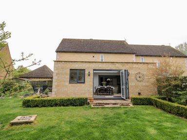 A backyard with lawn and trimmed hedges outside a stone house with open glass doors showing a dining table at Rosemead in Moreton-In-Marsh