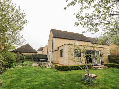 A backyard with a lawn and small tree next to a stone house with open doors at Rosemead in Moreton-In-Marsh