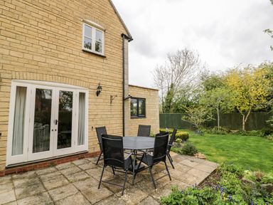 A patio area with a round table and six chairs outside a stone house with garden and trees at Rosemead in Moreton-In-Marsh
