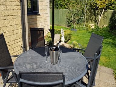 An outdoor patio area with a round metal table and four black mesh chairs a metal ice bucket with a bottle and two empty glasses on the table a garden with grass plants and a small statue behind the table at Rosemead in Moreton-In-Marsh