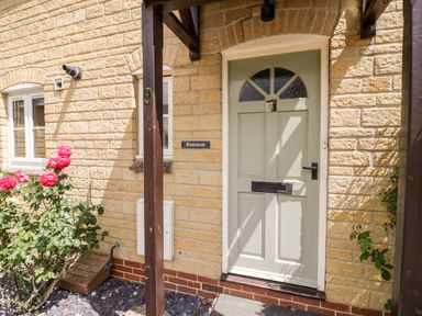 A front door with a porch and pink roses by the side of a stone house at Rosemead in Moreton-In-Marsh