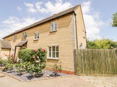 A stone house with white windows and a green fence with rose bushes and shrubs in the garden at Rosemead in Moreton-In-Marsh