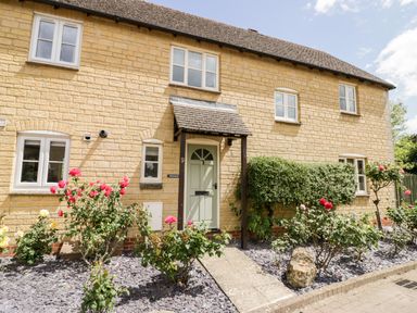 The exterior of a stone house with a green door and a garden with rose bushes and shrubs at Rosemead in Moreton-In-Marsh