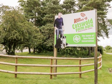 A large sign for Adam Hensons Cotswold Farm Park with a photo of a man and two dogs next to a wooden fence and trees at Rosemead in Moreton-In-Marsh