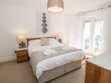 A bedroom with a double bed two bedside tables with lamps a dresser and glass doors with sheer curtains at Bay View in Trearddur Bay