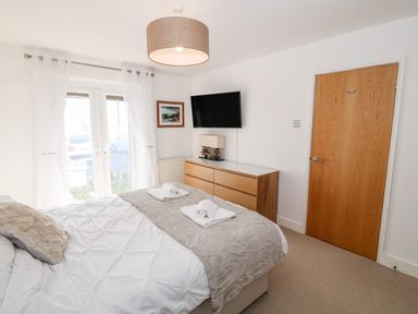 A bedroom with a bed with pillows and towels a wooden dresser a wall-mounted TV and a wooden door at Bay View in Trearddur Bay