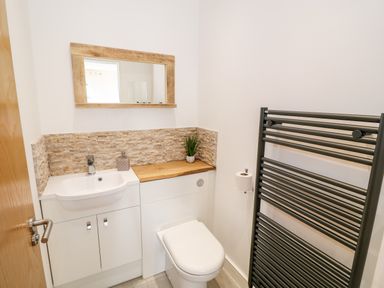 A small bathroom with a white sink and cabinets a toilet a wooden countertop a wall mirror and a black towel radiator at Bay View in Trearddur Bay