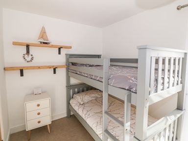A bedroom with a grey bunk bed next to a small white three-drawer cabinet and wooden shelves on the wall at Bay View in Trearddur Bay