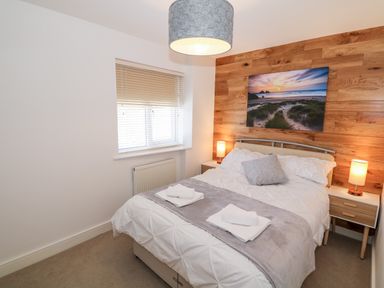 A bedroom with a double bed, bedside tables with lamps, a window with blinds, and a wooden accent wall with a beach photo at Bay View in Trearddur Bay
