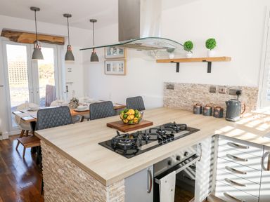 A kitchen with a wooden countertop, gas stove, kettle, jars, small plants on a shelf, and a dining table with chairs near glass doors at Bay View in Trearddur Bay