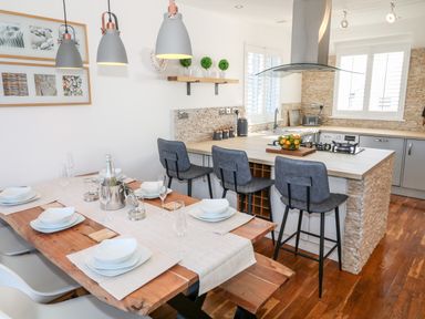 A kitchen with a wooden dining table set with white dishes and glasses and a kitchen island with three gray bar stools at Bay View in Trearddur Bay