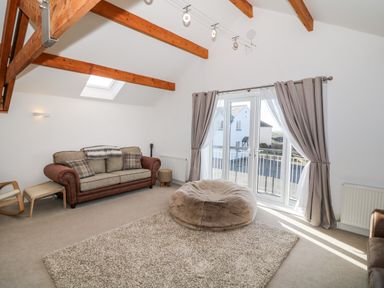 A living room with a sofa a bean bag a woven ottoman a guitar and large windows with curtains at Bay View in Trearddur Bay