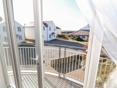 A view through glass doors to a small curved balcony with metal railing overlooking a street with white houses and a distant sea view at Bay View in Trearddur Bay