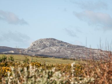 A rocky hill behind a grassy field with bushes in the foreground at Bay View in Trearddur Bay