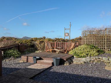 An outdoor wooden deck with two wooden chairs and a small table surrounded by plants and a wooden fence at Bay View in Trearddur Bay