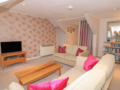 A living room with beige sofas and pink cushions a wooden coffee table a tv on a wooden stand floral wallpaper and bookshelves at Manor Studio in Dartmouth
