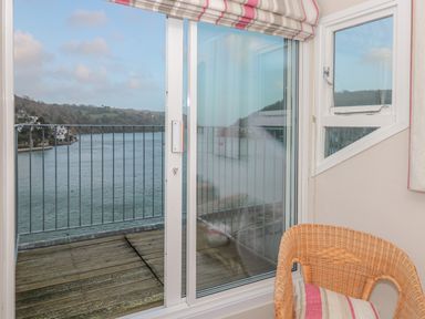 A sliding glass door opening to a balcony overlooking a river with hills in the background and a wicker chair with a striped cushion inside the room at Manor Studio in Dartmouth