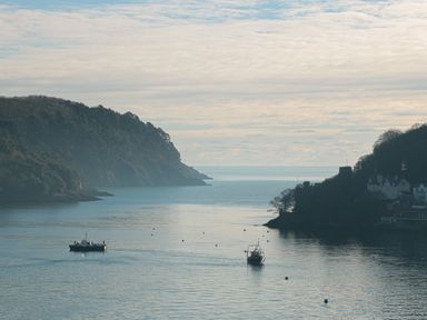 A coastal scene with two boats on calm water between forested hills and houses at Manor Studio in Dartmouth