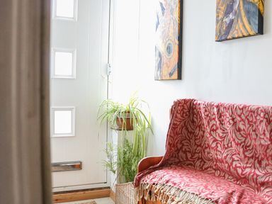 A hallway with a wooden bench covered with a patterned red blanket two paintings on a white wall and green plants near a white door with square windows at 21 Florence Road in Pakefield
