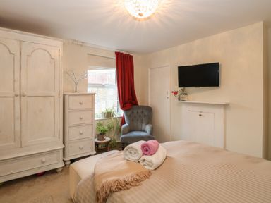 A bedroom with a bed, rolled towels, a chair, wardrobe, chest of drawers, a window with red curtains and a wall-mounted television at 21 Florence Road in Pakefield