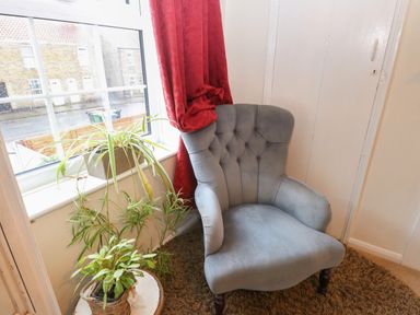 A gray upholstered armchair near a window with red curtains and two potted plants at 21 Florence Road in Pakefield