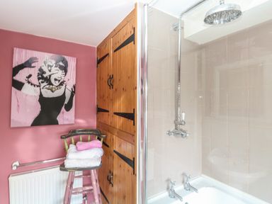 A bathroom with a bathtub shower and wooden cabinets next to a pink wall with a painting and towels on a chair at 21 Florence Road in Pakefield