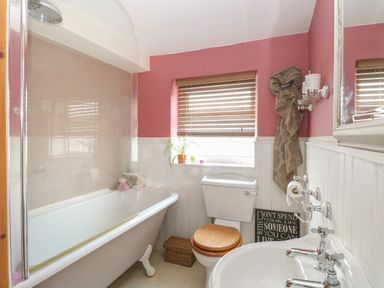 A bathroom with a bathtub with shower, a toilet with wooden seat, a sink, a window with blinds and a plant on the sill at 21 Florence Road in Pakefield