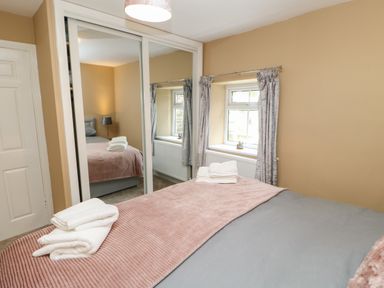 A bedroom with a bed covered by a grey duvet and pink blanket with folded towels on top a window with grey curtains and a wardrobe with mirrored sliding doors at Crag View in West Woodburn