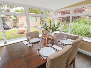 A dining table set for four with plates glasses and a bottle of wine in a room with large windows overlooking a garden at Crag View in West Woodburn