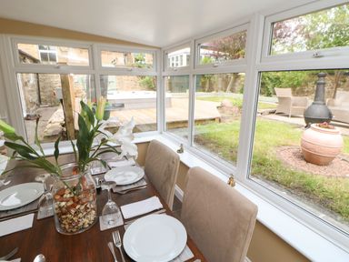 A dining table with plates glasses and a vase with flowers next to large windows showing a garden at Crag View in West Woodburn