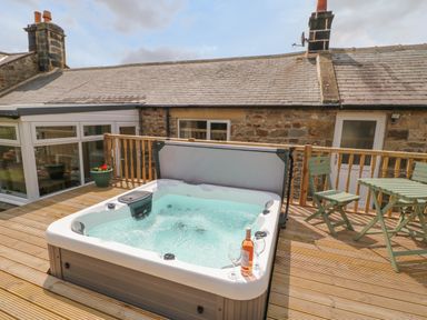 A hot tub on a wooden deck outside a stone house with a table and chairs at Crag View in West Woodburn