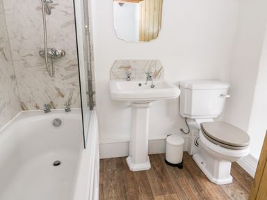 A bathroom with a bathtub shower sink toilet and a small trash bin on a wooden floor