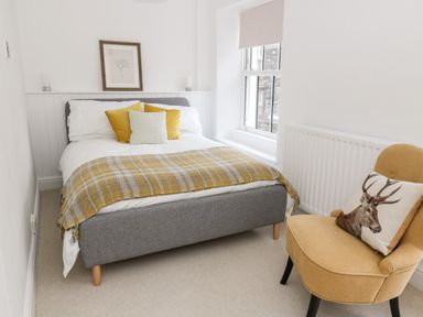 A bedroom with a bed with yellow and white pillows and a blanket and a yellow chair with a deer pillow at Pine Cottage in Sedbergh