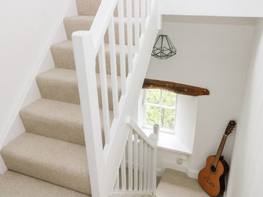 A staircase with beige carpet a window with a wooden beam and a guitar in the corner at Pine Cottage in Sedbergh
