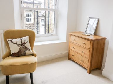A yellow chair with a deer print pillow next to a window and a wooden dresser with a framed picture on top in a room