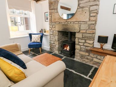 A living room with a stone fireplace and wood stove a beige sofa with pillows a blue chair and a wooden table at Pine Cottage in Sedbergh