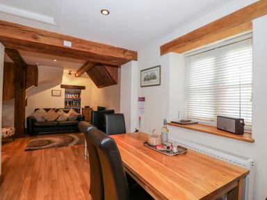 A dining area with a wooden table and chairs next to a window with blinds and a living area with a sofa and wooden beams at Chapel Cottage in Leonard Stanley near Kings Stanley