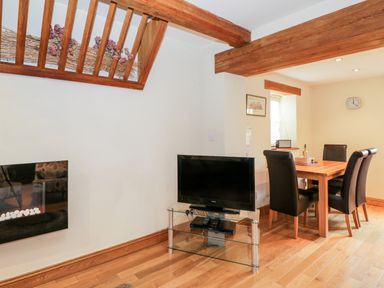 A dining area with a wooden table and four black chairs next to a glass TV stand with a television at Chapel Cottage in Leonard Stanley near Kings Stanley
