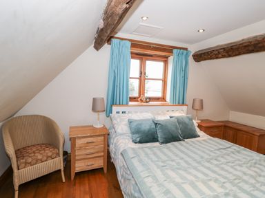 A bedroom with a bed pillows bedside tables lamps a wicker chair wooden beams and blue curtains at Chapel Cottage Leonard Stanley near Kings Stanley