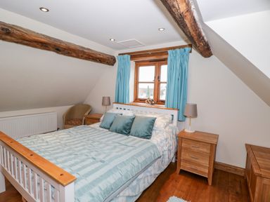A bedroom with a wooden bed frame blue bedding two wooden bedside tables with lamps a window with blue curtains and ceiling beams at Chapel Cottage in Leonard Stanley near Kings Stanley