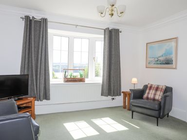 A living room with armchairs a TV on a wooden stand and a window with grey curtains and plants at Sea View Cottage in Menai Bridge