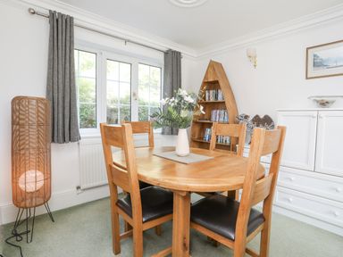A dining room with a round wooden table and four chairs a wooden bookshelf shaped like a boat and a wicker floor lamp at Sea View Cottage in Menai Bridge