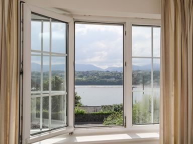 A window with beige curtains looking out over water and hills at Sea View Cottage in Menai Bridge